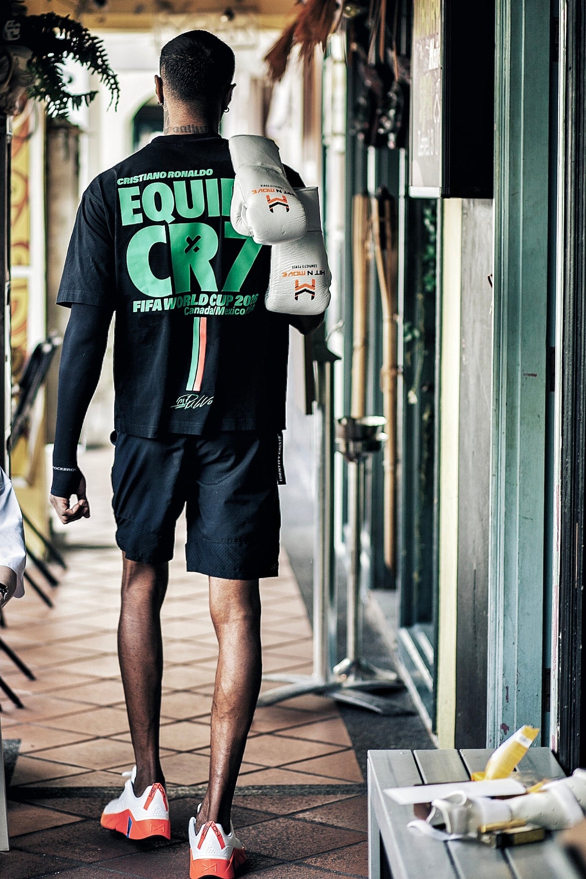A boxer seen from behind wearing a black Equipa CR7 FIFA World Cup 2026 oversized boxy t-shirt with a large green and white graphic on the back, black athletic shorts, and white and orange sneakers, while carrying white boxing gloves over his shoulder in an outdoor corridor.