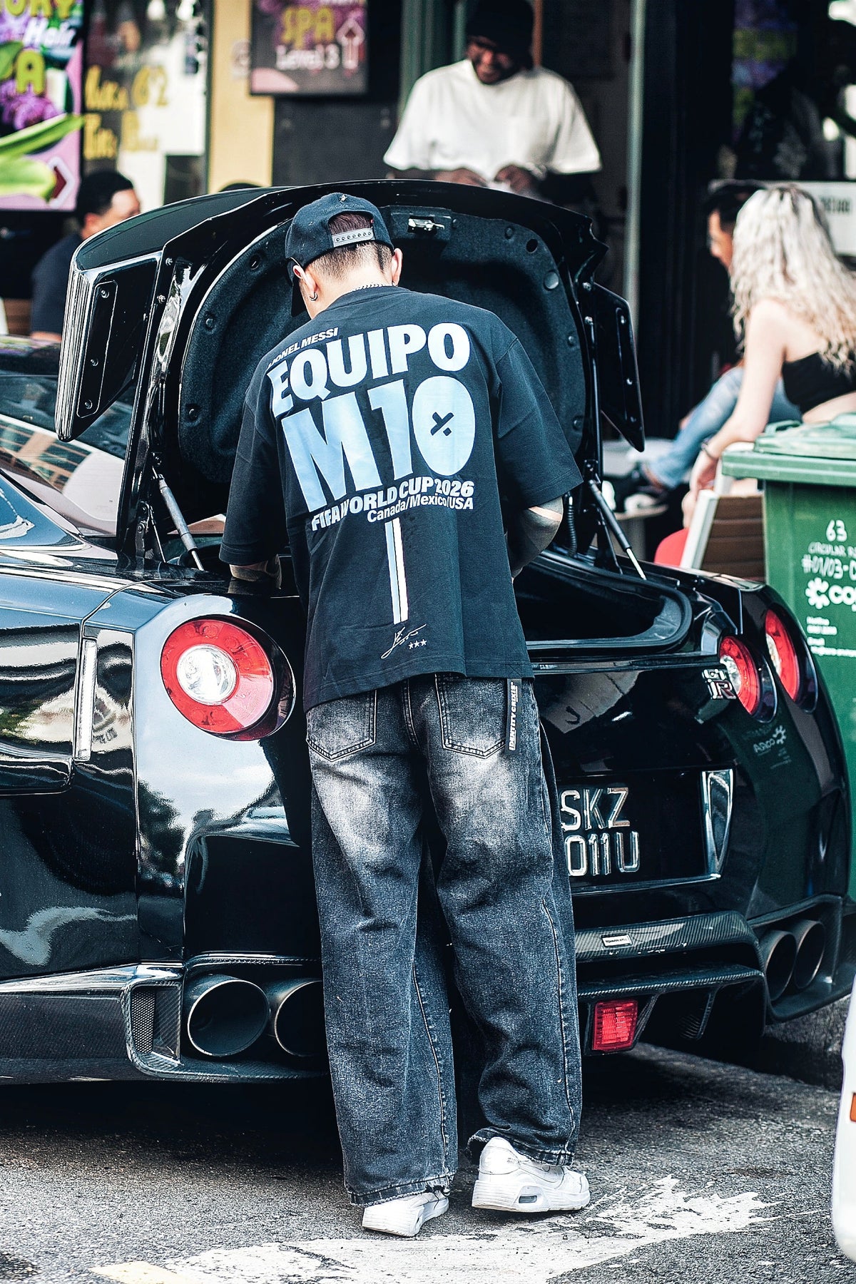 A man stands at the open trunk of a black Nissan GTR car in a street setting, wearing a black graphic T-shirt with 'EQUIPO M10' printed on the back, dark jeans, and white sneakers. The background shows a tattoo parlor storefront with people inside.