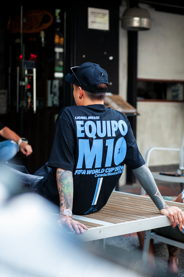 A man wearing a black Lionel Messi 'Equipo M10' FIFA World Cup 20226 t-shirt and a matching black cap, sitting on a wooden bench in an outdoor cafe setting.