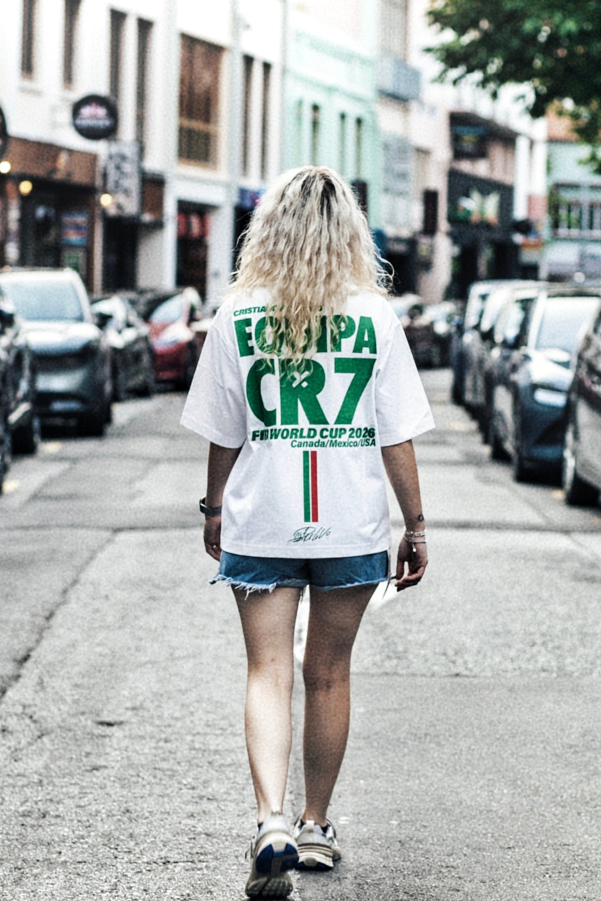 A woman walking down a city street away from the camera, wearing a white t-shirt with green and red text on the back that reads 'EQUIPA CR7' and denim shorts.